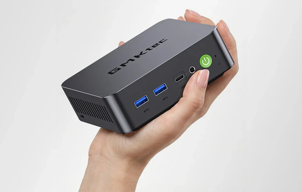 Rankoje laikomas GMKtec mini kompiuteris, matomas priekinis skydelis su maitinimo mygtuku, USB prievadais, USB-C prievadu ir garso jungtimi.